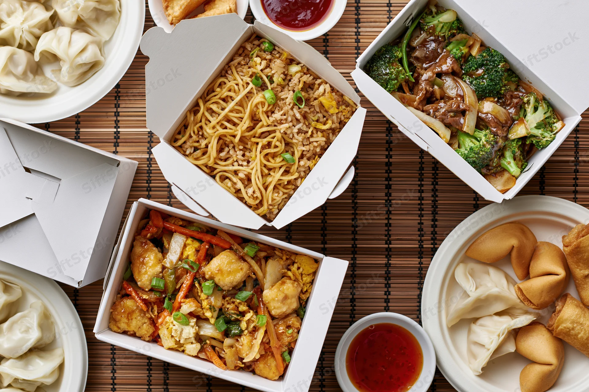 Overhead flat lay of multiple chinese takeout boxes filled with noodles, stir-fried vegetables and meat, dumplings, and sauces on a bamboo mat