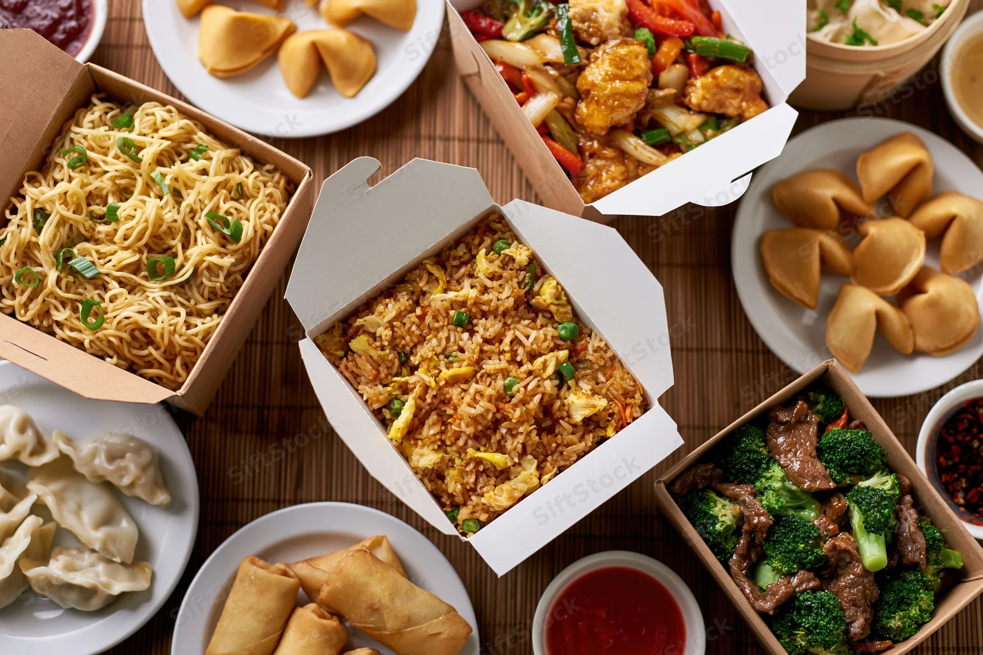 Overhead flat lay of multiple chinese takeout boxes and plates with fried rice, noodles, beef and broccoli, fortune cookies, dumplings, and spring rolls on a bamboo mat