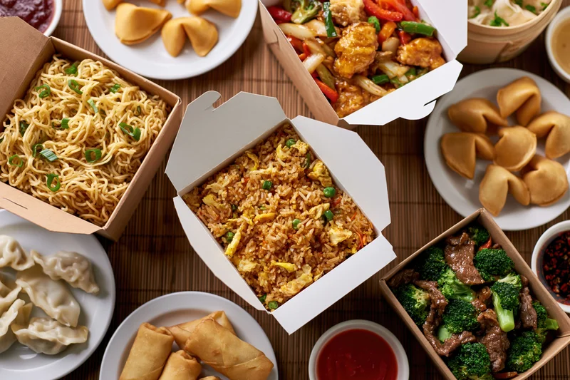 Overhead flat lay of multiple chinese takeout boxes and plates with fried rice, noodles, beef and broccoli, fortune cookies, dumplings, and spring rolls on a bamboo mat