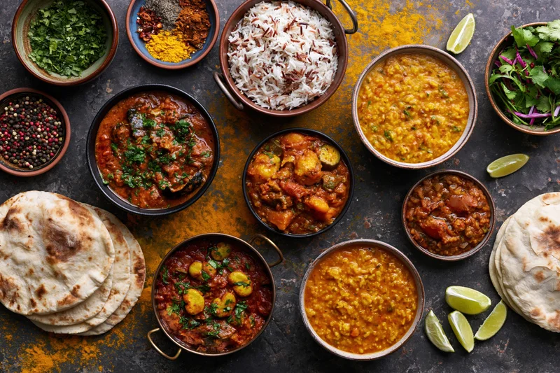 Overhead flat lay of multiple indian curry dishes rice dal and naan on dark textured surface with spices and lime wedges