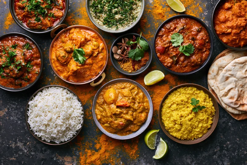 Overhead flat lay of multiple indian curry dishes rice dal and naan on dark textured surface
