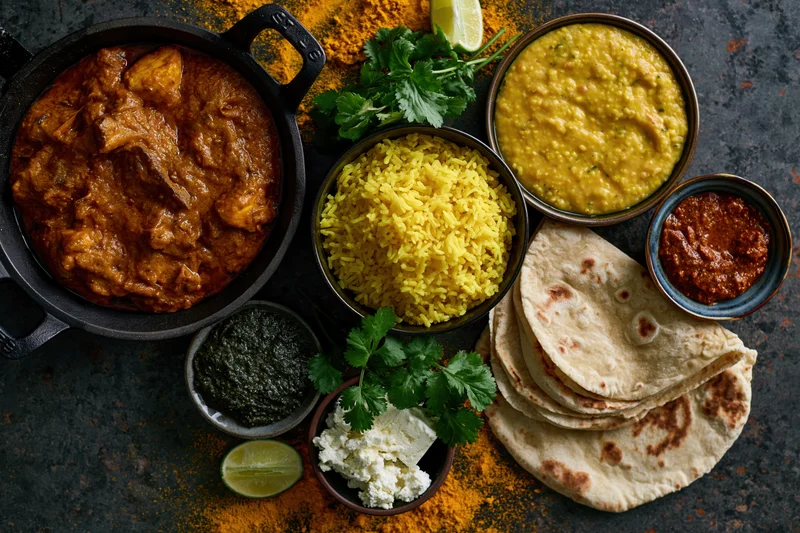 Overhead flat lay of indian dishes including curry, rice, naan, dal, chutney, yogurt, and cilantro on dark textured surface