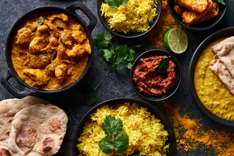 Overhead flat lay of multiple indian dishes arranged on dark surface with curry, rice, naan, chutney, lime, and cilantro