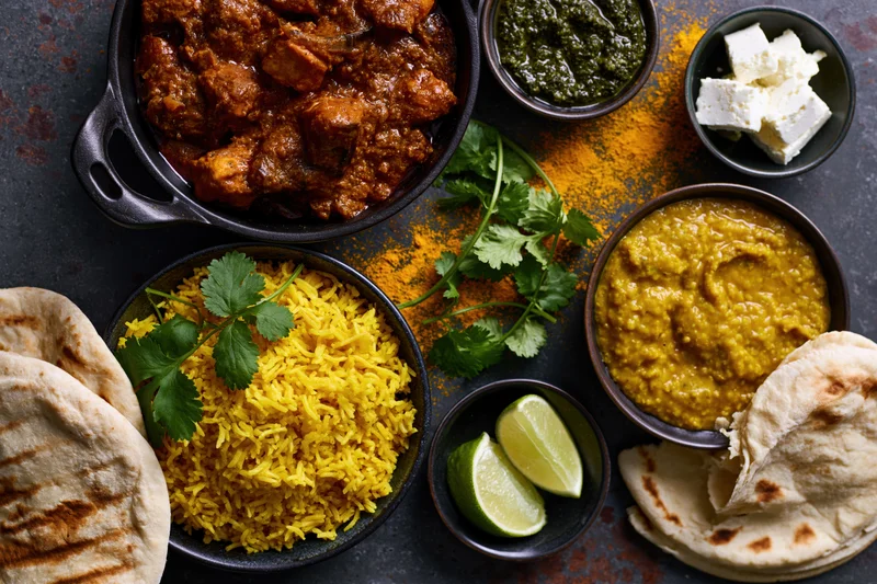 Overhead flat lay of indian dishes including curry, yellow rice, dal, naan, cilantro, lime, and turmeric spice on dark textured surface
