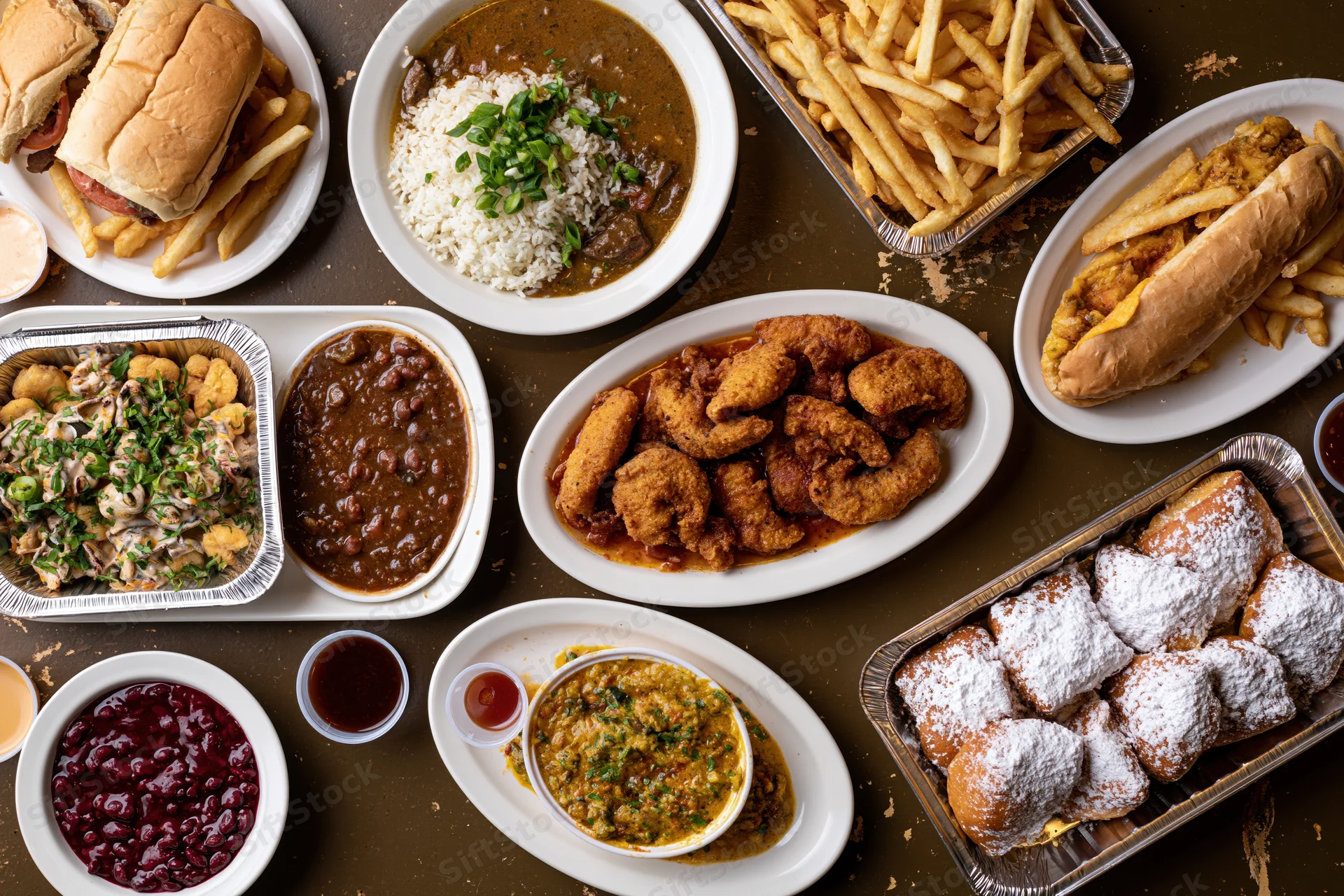 Overhead flat lay of multiple southern cajun restaurant dishes arranged on a table with aluminum trays and white plates, featuring gumbo, fried chicken, po'boys, beignets, and side dishes