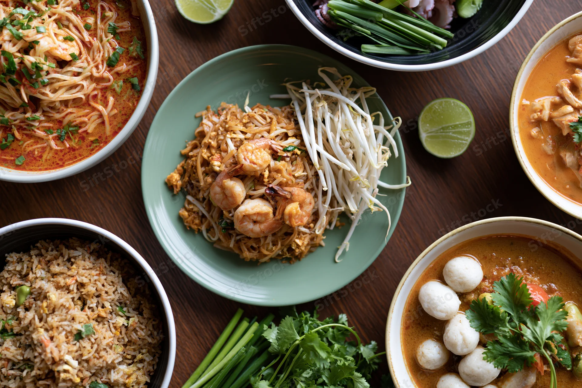 Overhead flat lay of multiple thai dishes arranged on a wooden table with garnishes and sauces