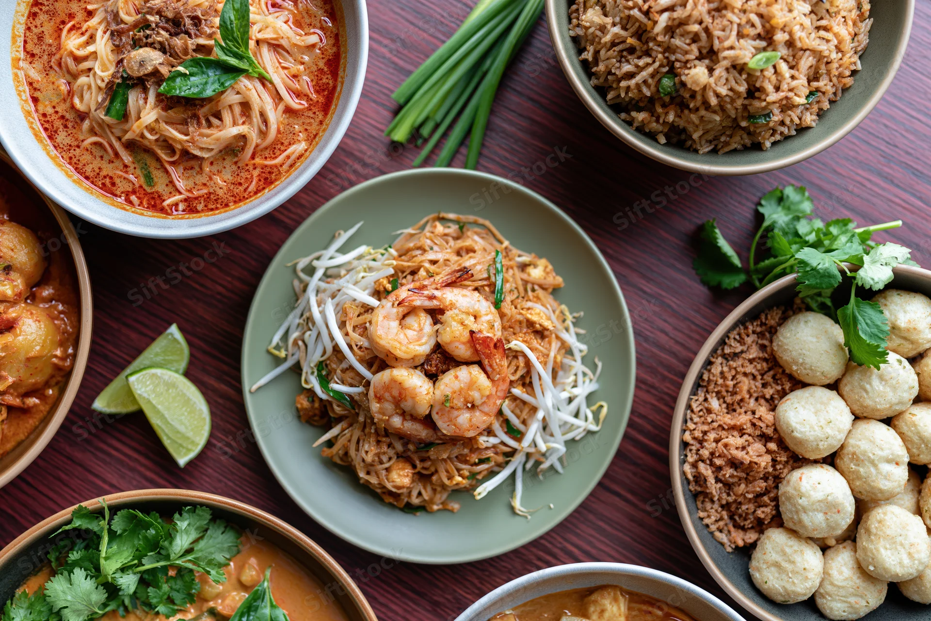 Overhead flat lay of multiple thai dishes arranged on a dark wooden table with bowls of noodles, rice, curry, and garnishes