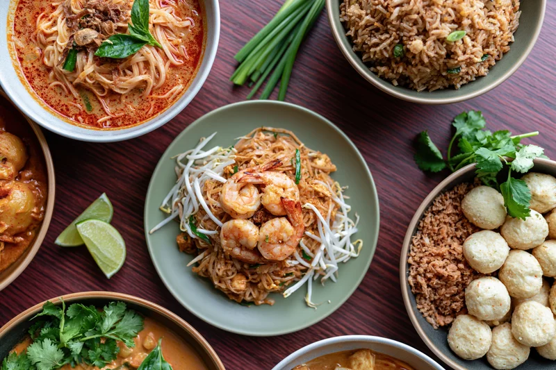 Overhead flat lay of multiple thai dishes arranged on a dark wooden table with bowls of noodles, rice, curry, and garnishes
