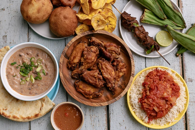 Overhead flat lay of multiple West African dishes arranged on a wooden table with rice, stew, fried chicken, plantain, and skewers
