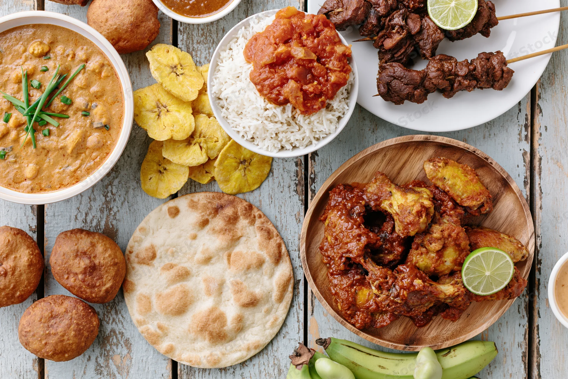 Overhead flat lay of various West African dishes arranged on a wooden table including rice, curry, skewered meat, plantain, and flatbread