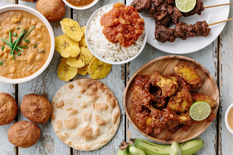Overhead flat lay of various West African dishes arranged on a wooden table including rice, curry, skewered meat, plantain, and flatbread