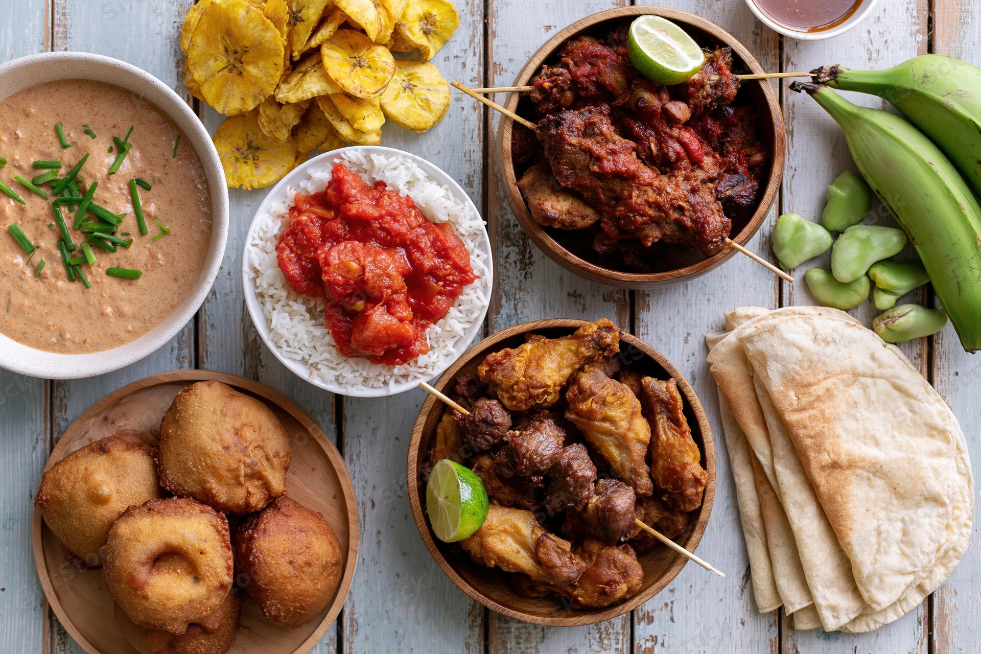 Overhead flat lay of multiple West African dishes arranged on a wooden table with bowls of stew, rice, fried snacks, plantains, and flatbread
