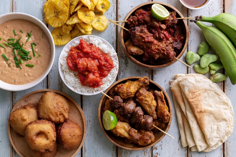 Overhead flat lay of multiple West African dishes arranged on a wooden table with bowls of stew, rice, fried snacks, plantains, and flatbread