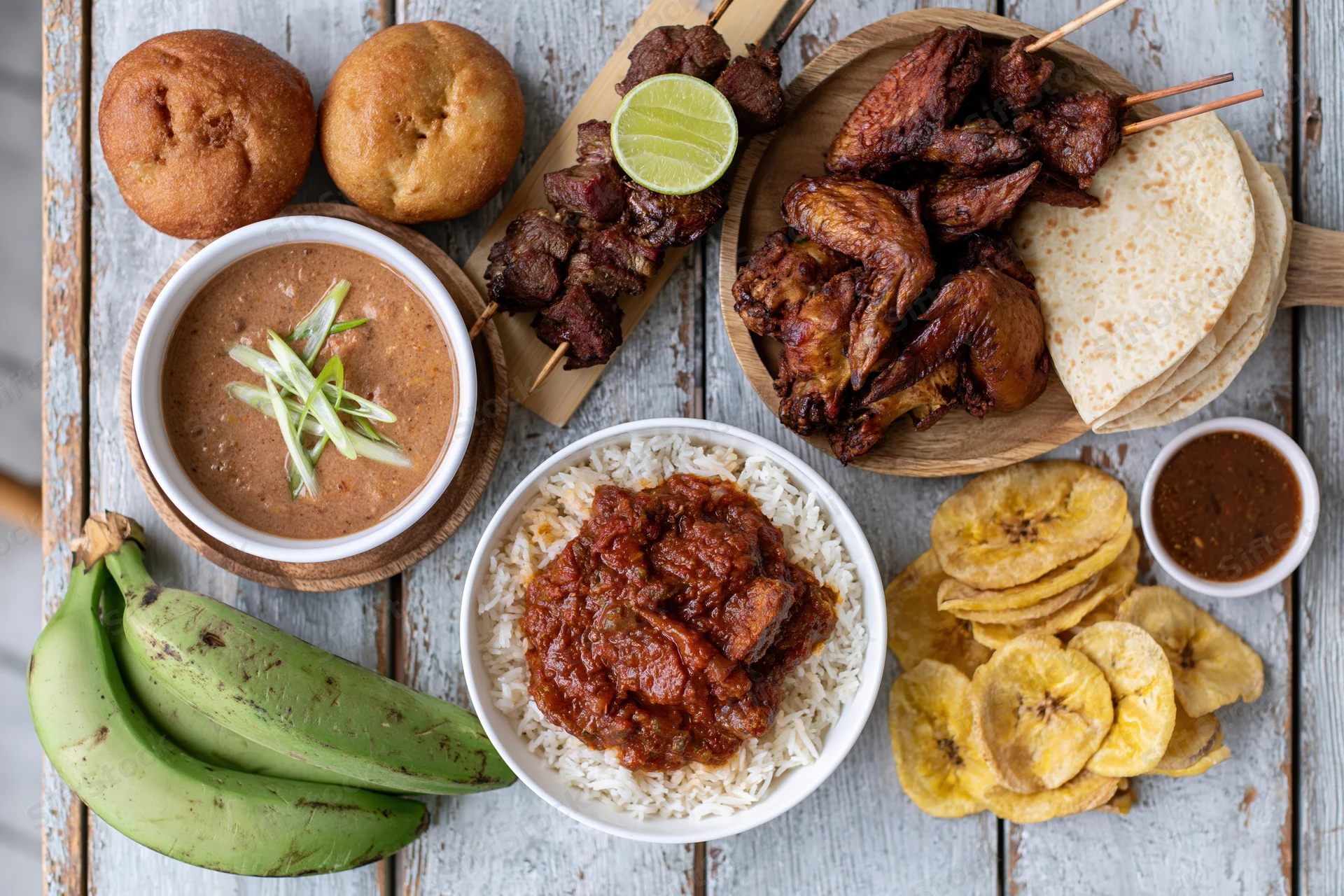 Overhead flat lay of west african dishes including rice with stew, grilled meats on skewers, plantain chips, buns, and sauces arranged on a wooden table