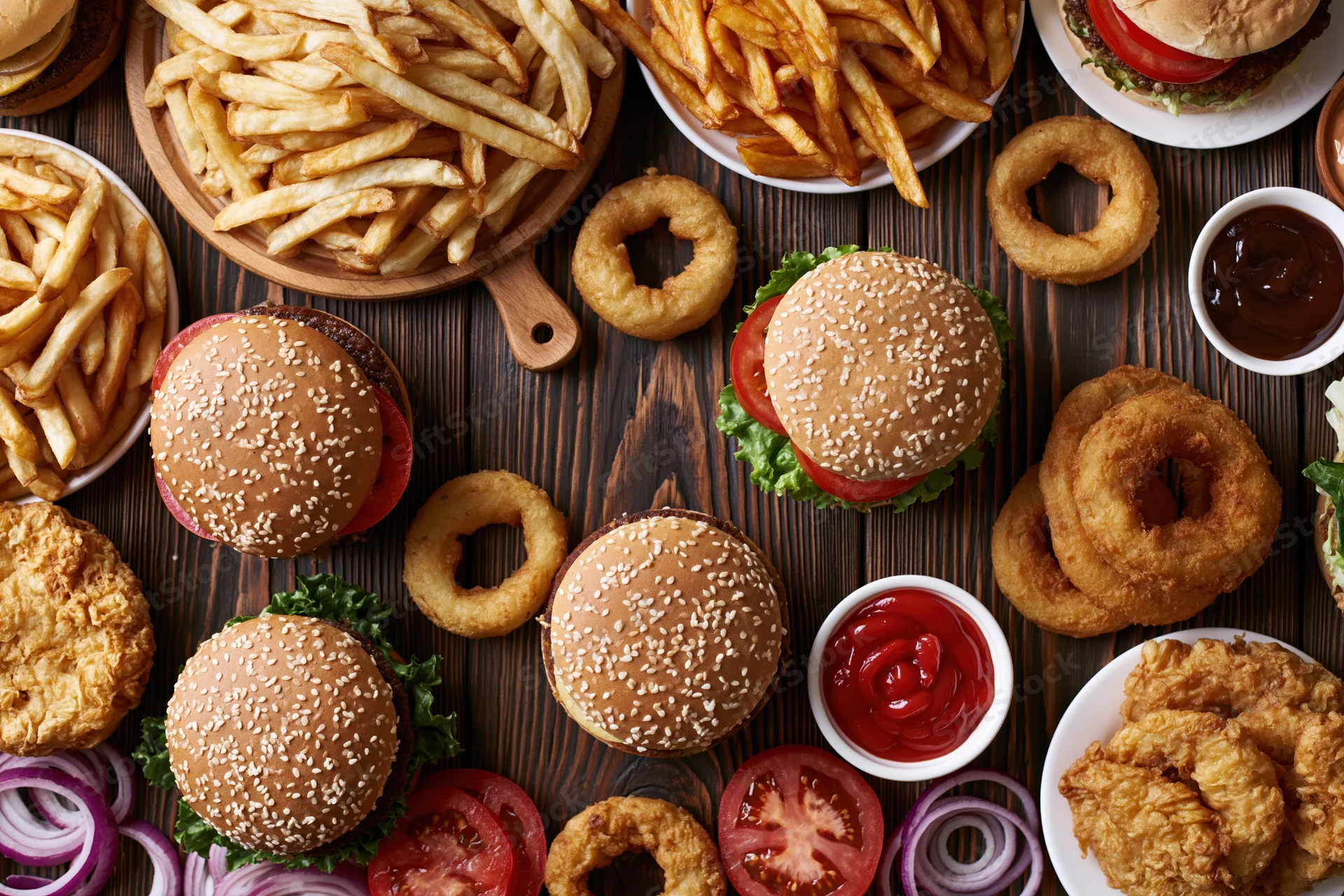 Overhead flat lay of multiple burgers, french fries, onion rings, and condiments on a dark wooden table
