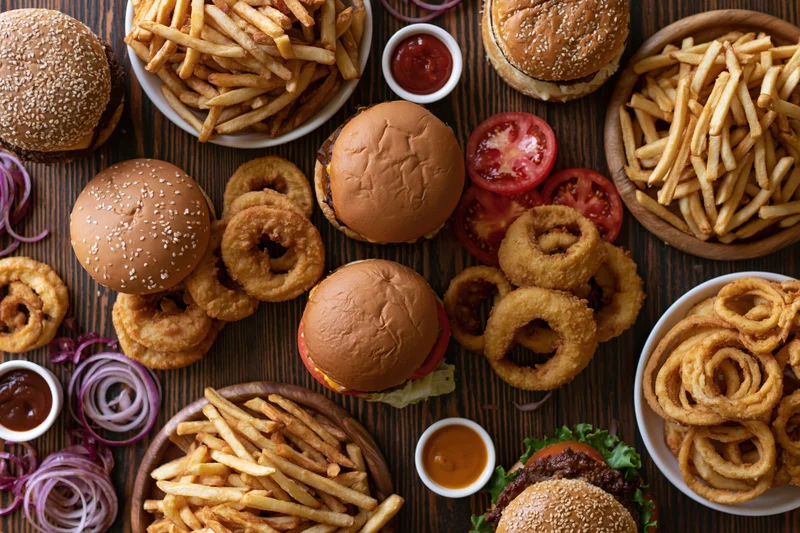 Multiple burgers and fries with onion rings arranged on a wooden table in overhead flat lay view