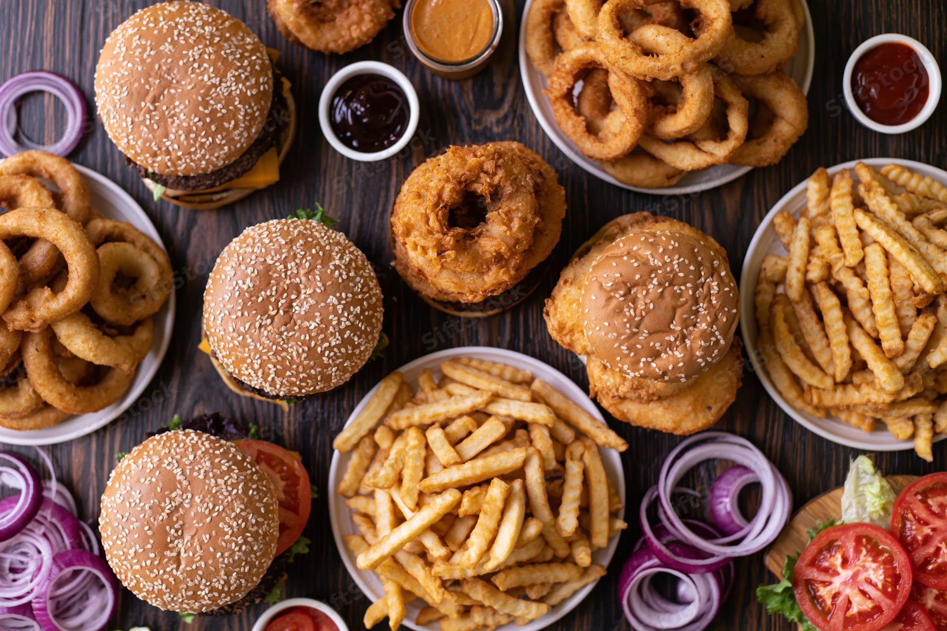 Multiple burgers and fries with onion rings and condiments arranged on a dark wooden table in overhead flat lay view