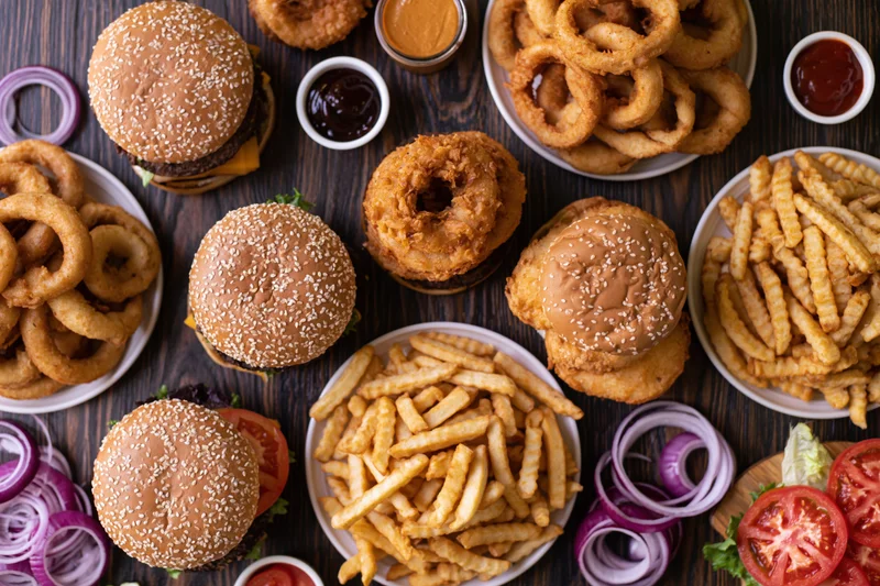 Multiple burgers and fries with onion rings and condiments arranged on a dark wooden table in overhead flat lay view
