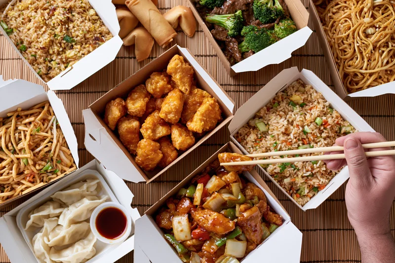 Overhead view of multiple open chinese takeout boxes arranged on a bamboo mat with a hand using chopsticks to pick up food
