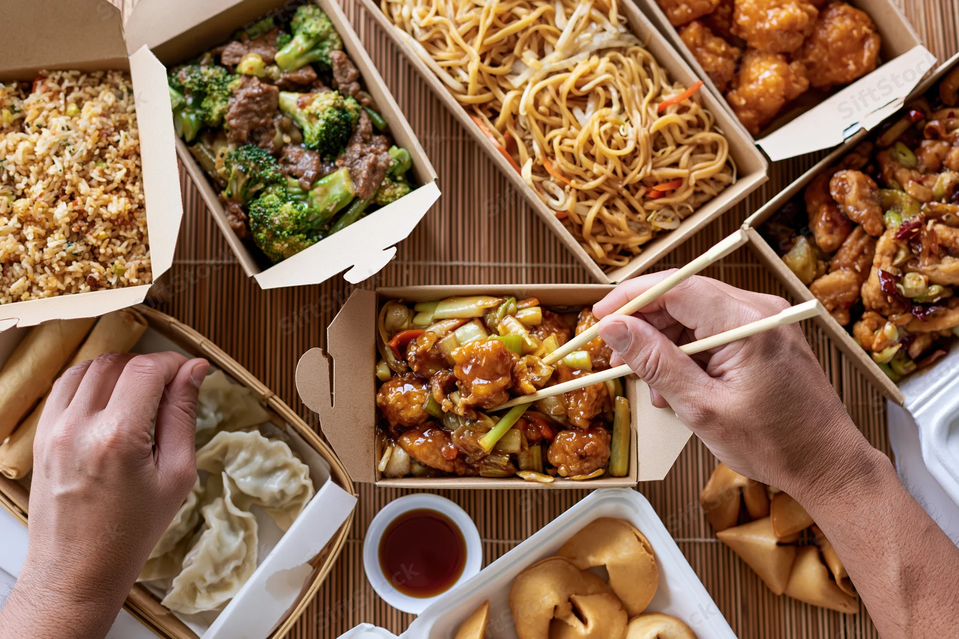 Overhead view of multiple open chinese takeout boxes with hands using chopsticks on a bamboo mat