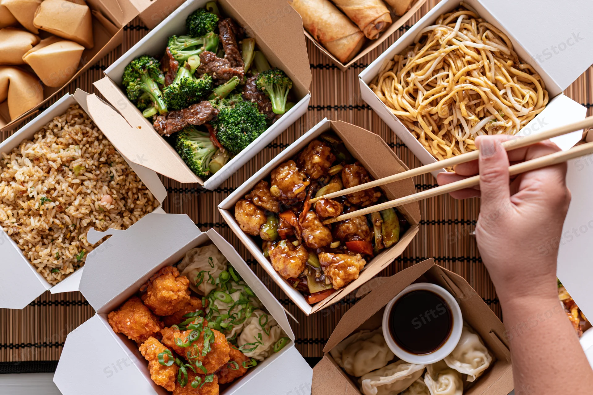 Overhead view of multiple open chinese takeout boxes arranged on a bamboo mat with a hand using chopsticks to pick food