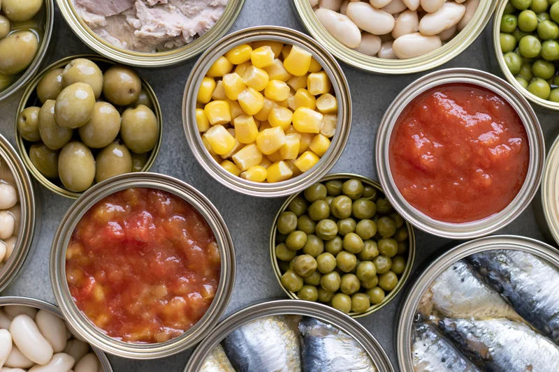 Overhead flat lay of multiple open tin cans filled with various canned foods arranged on a dark surface