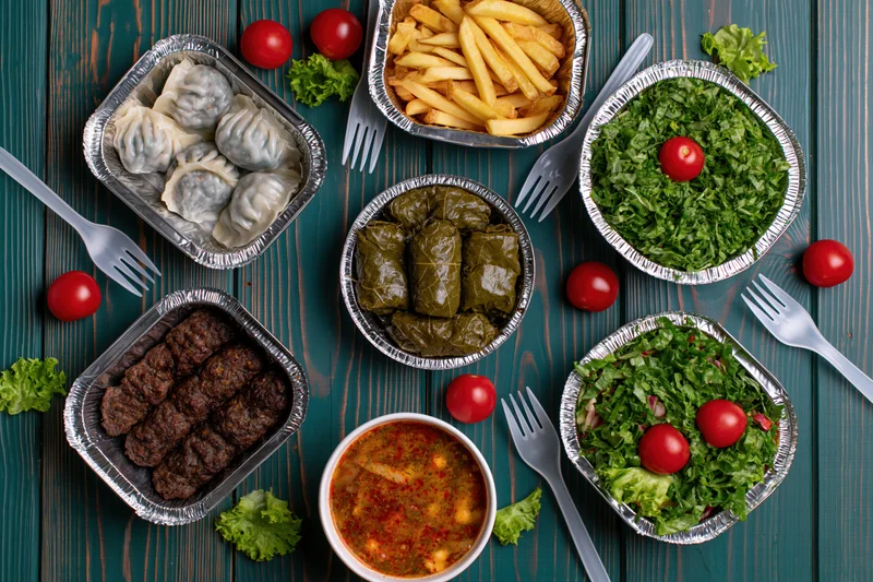 Overhead flat lay of multiple takeout dishes arranged on a wooden table with foil containers holding dumplings, meatballs, stuffed grape leaves, salads, fries, and soup, garnished with tomatoes and lettuce, surrounded by plastic forks
