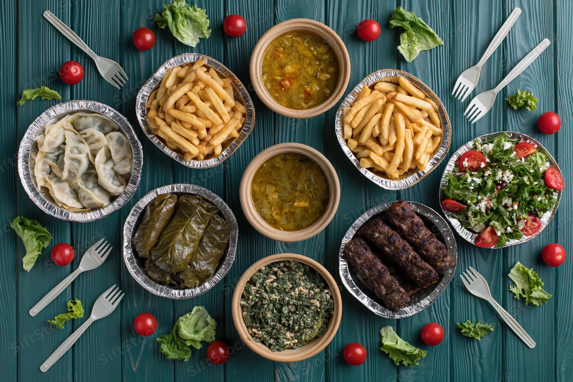 Overhead flat lay of multiple takeout dishes arranged on a blue wooden table with forks and garnishes
