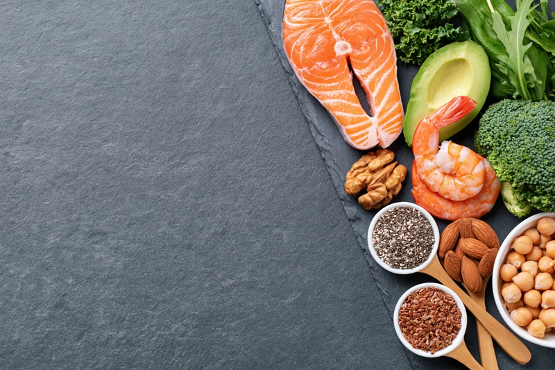 Overhead flat lay of salmon, shrimp, avocado, broccoli, nuts, seeds, and legumes arranged on slate with copyspace