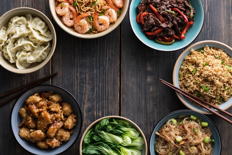 Seven asian dishes arranged in a circle on a dark wooden table with chopsticks and bowls