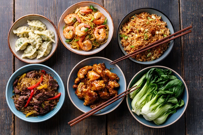 Seven asian dishes arranged in a circle on a wooden table with chopsticks and ceramic bowls