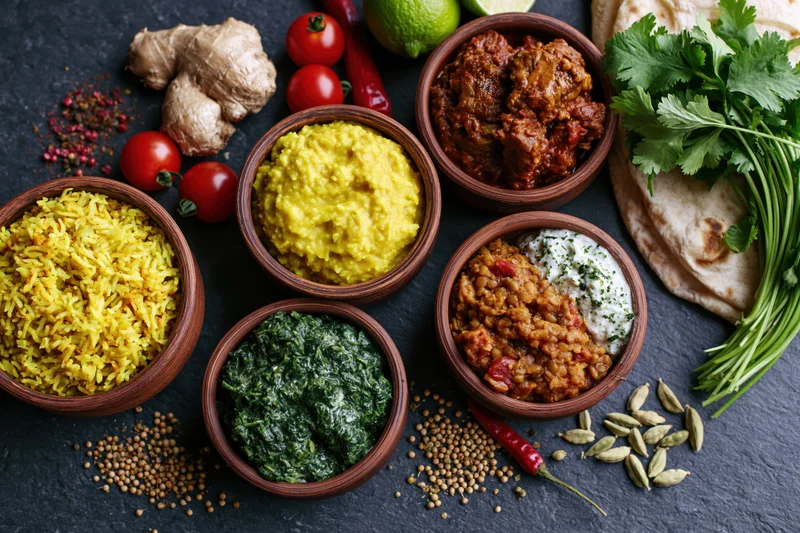 Overhead flat lay of indian dishes in clay bowls arranged on dark slate surface with fresh herbs and spices