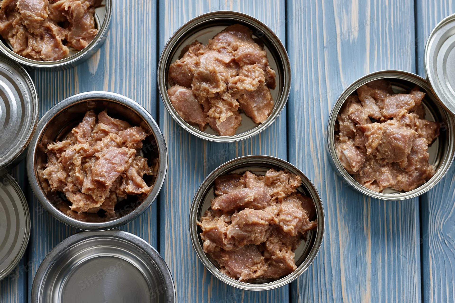 Open metal cans of chunky pet food arranged on a blue wooden surface