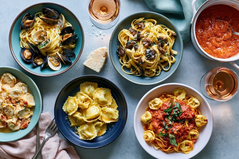 Overhead flat lay of six pasta dishes arranged on a gray table with wine glasses and napkins