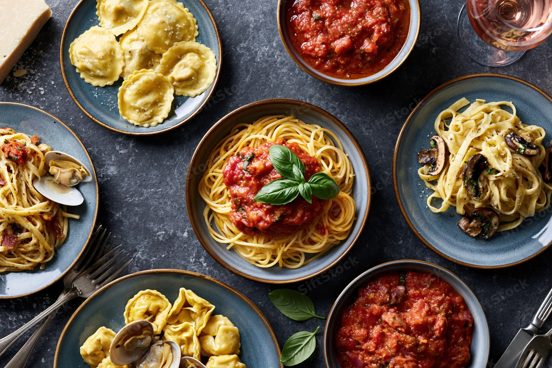 Overhead flat lay of six pasta dishes arranged on dark surface with sauce, basil, mushrooms, clams, cheese, and wine