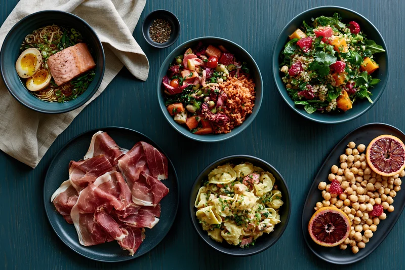 Overhead flat lay of six varied meal bowls and plates arranged on a dark wooden table