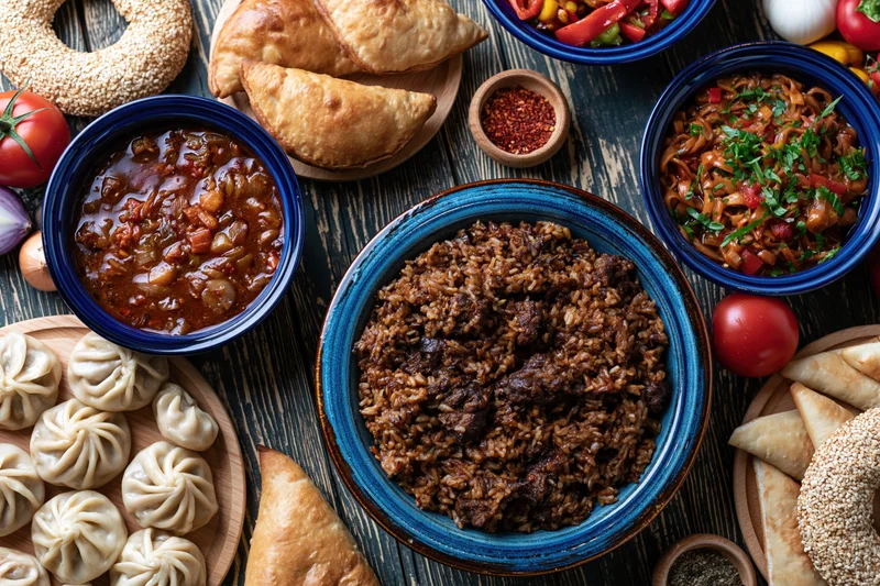 Overhead flat lay of central asian plov with meat and rice served with dumplings, samosas, and vegetables on a wooden table