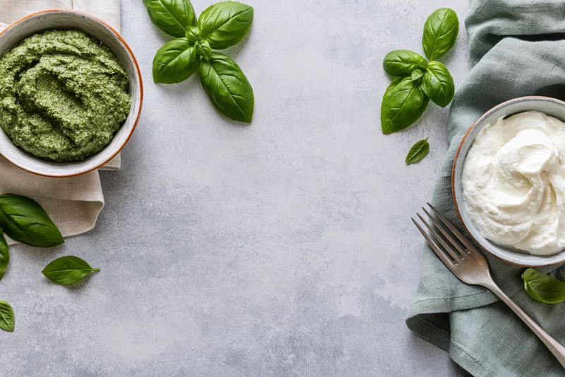 Two ceramic bowls with green pesto and white ricotta cheese arranged on a gray surface with fresh basil leaves and a fork nearby