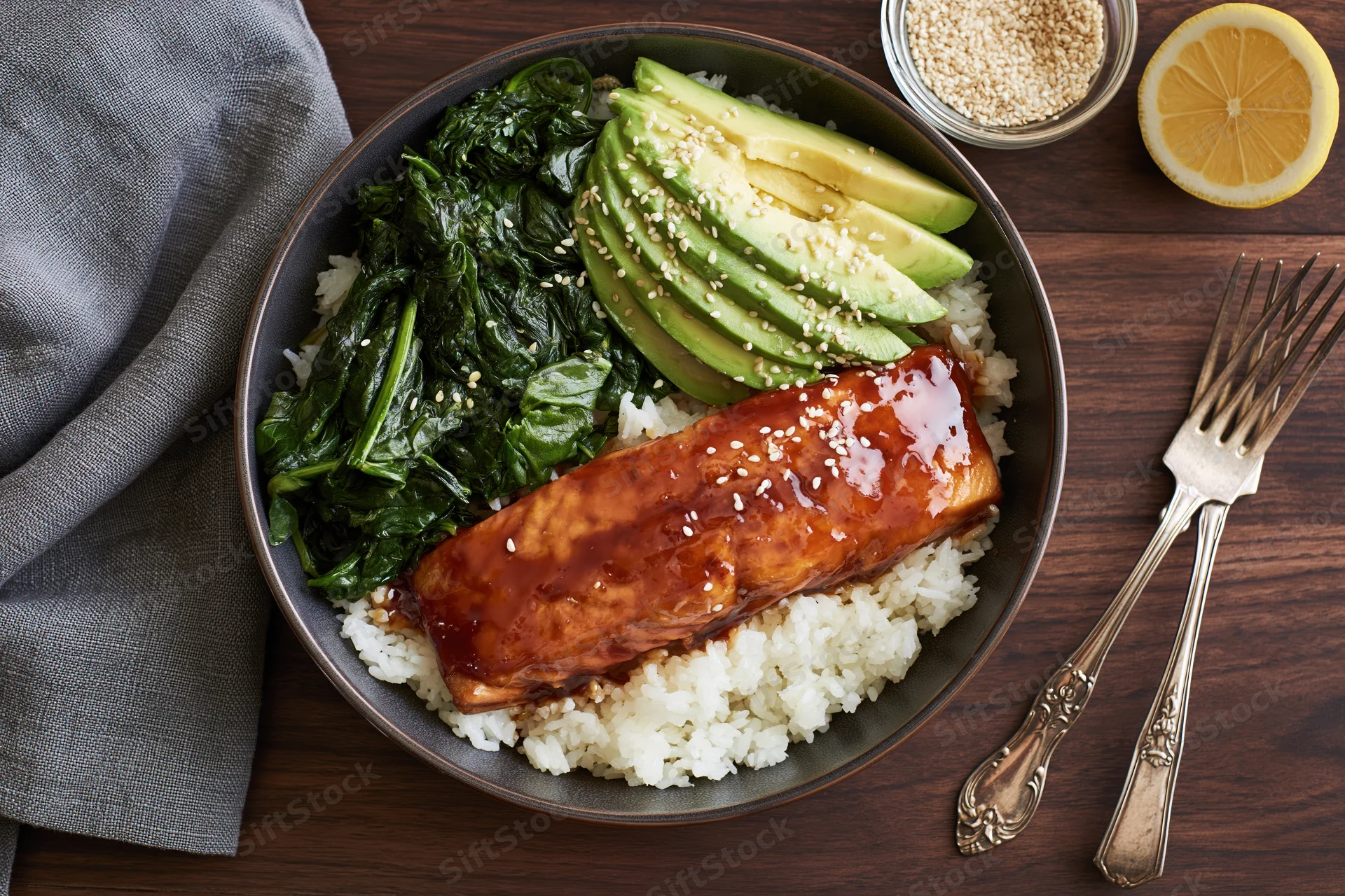 Salmon fillet with teriyaki glaze and sesame seeds served over white rice with avocado and spinach in a dark ceramic bowl on wooden table with fork and lemon slice