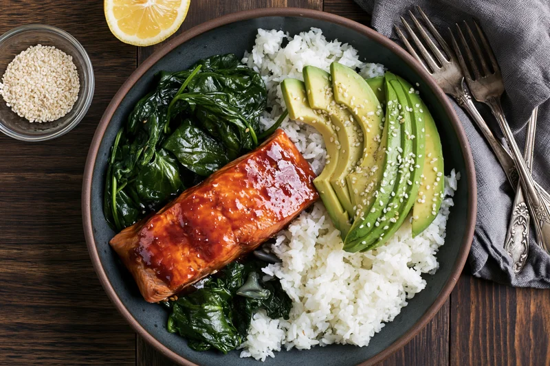 Glazed salmon with rice, avocado, and spinach in a dark ceramic bowl on a wooden table