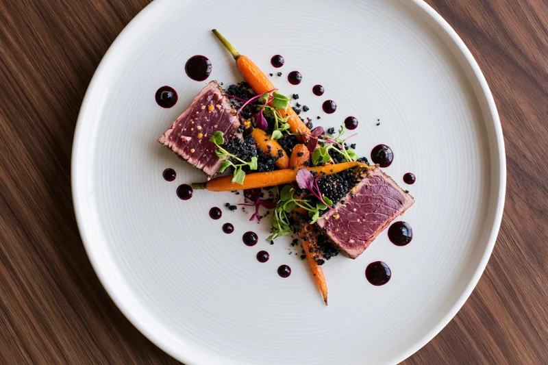 Seared tuna medallions with carrots and microgreens on a white plate over dark wood table