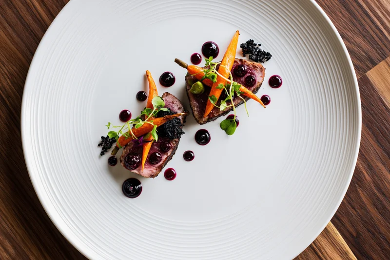 Two seared tuna medallions with orange carrots and berry sauce on a white plate on a wooden table