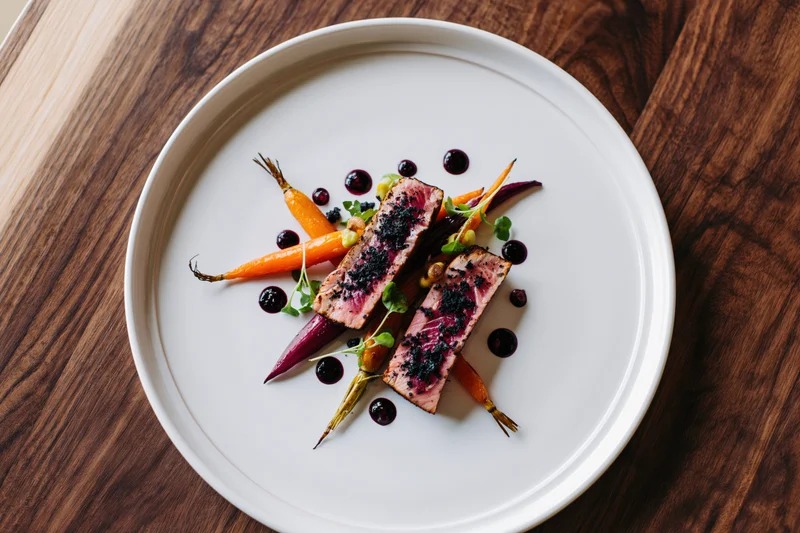 Two seared tuna medallions with roasted carrots and berry sauce on a white plate on a wooden table