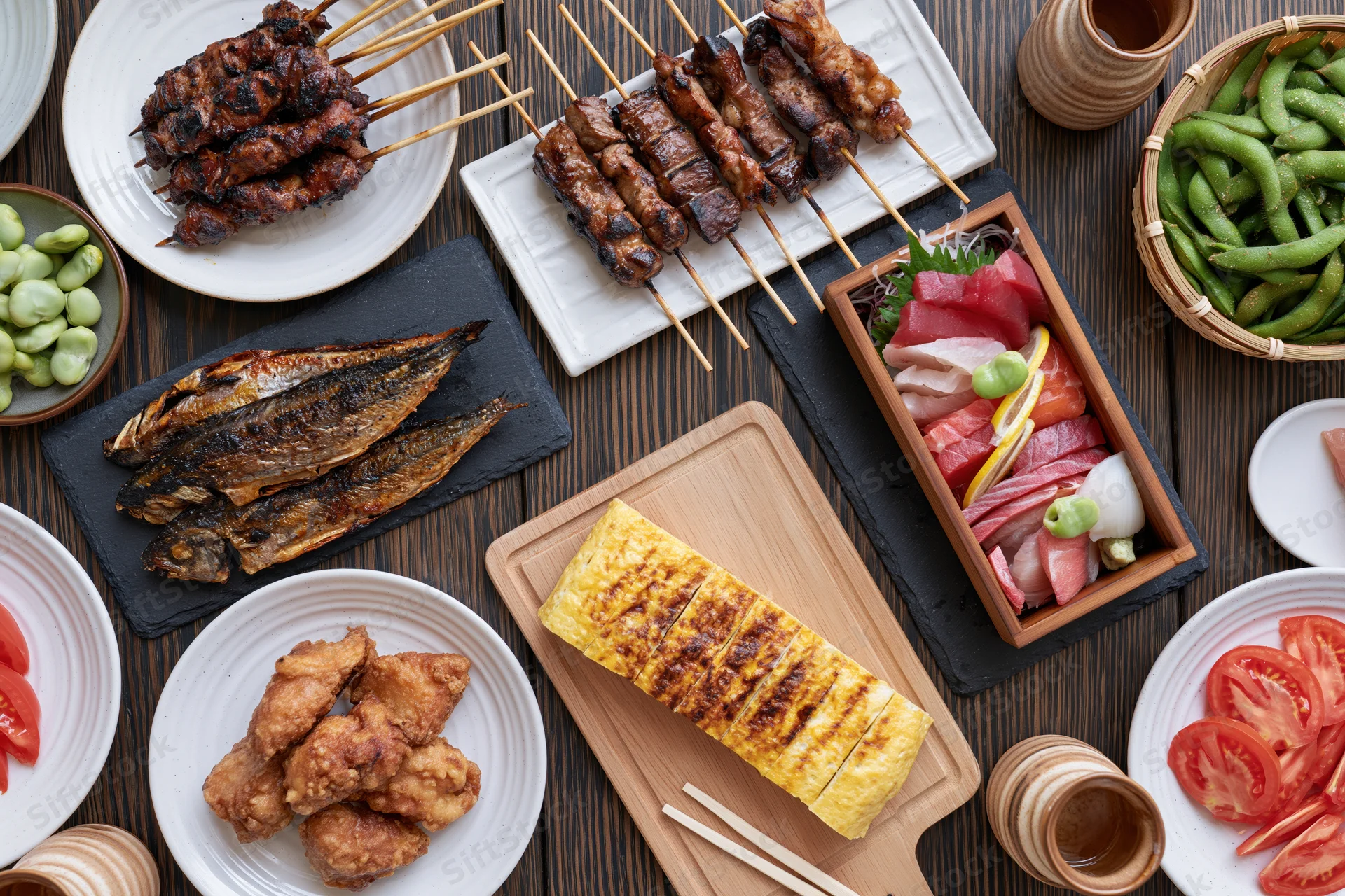 Overhead view of Japanese izakaya small dishes arranged on a wooden table with skewers, grilled fish, sashimi, edamame, and tamagoyaki