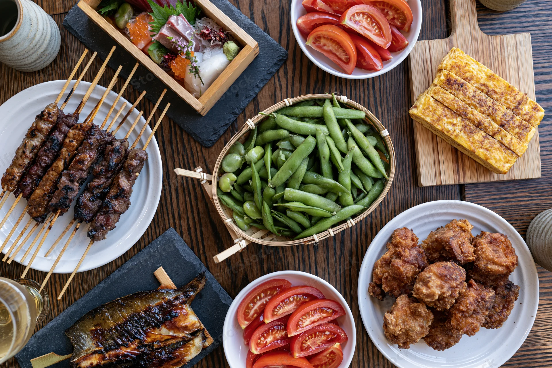 Overhead view of Japanese izakaya small dishes arranged on a dark wooden table with skewers, edamame, tempura, sushi, and grilled fish