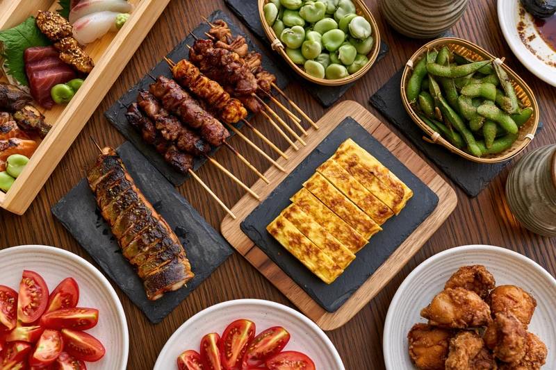 Overhead view of Japanese izakaya small dishes arranged on a wooden table with skewers, grilled salmon, tamagoyaki, edamame, and fried chicken