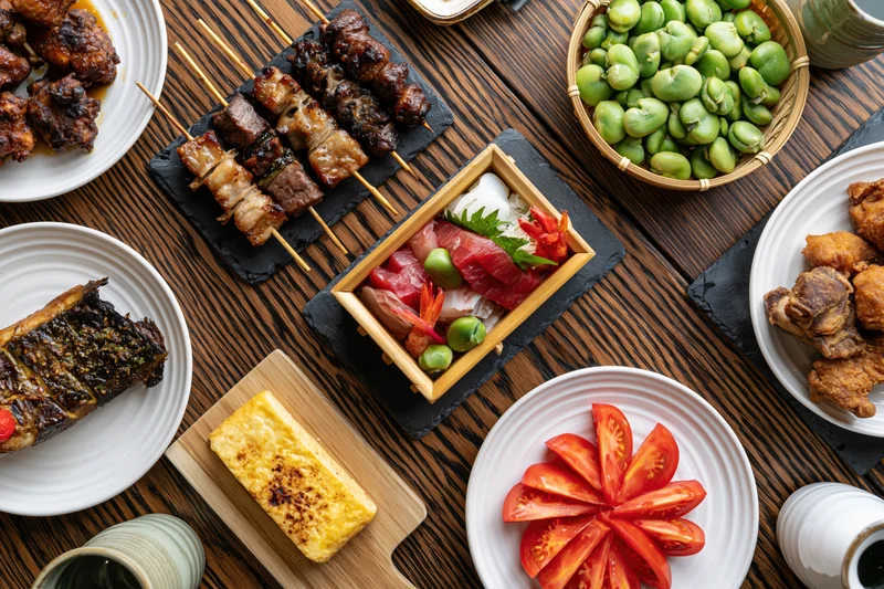 Overhead view of Japanese izakaya small dishes arranged on a wooden table with skewers, sashimi, edamame, and tempura