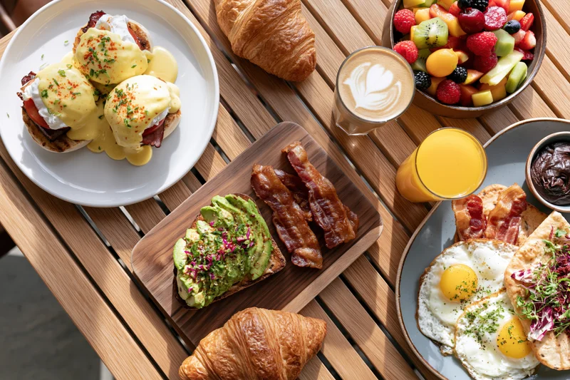 Overhead view of a brunch spread with eggs benedict, avocado toast, bacon, croissants, fruit salad, coffee and orange juice on a wooden table