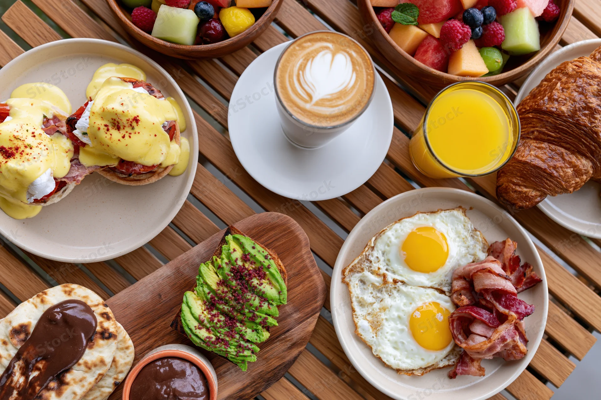 Overhead view of a brunch table with eggs benedict, avocado toast, croissant, bacon, fried eggs, orange juice, latte, and fruit bowls on a wooden table