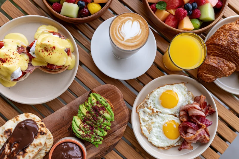 Overhead view of a brunch table with eggs benedict, avocado toast, croissant, bacon, fried eggs, orange juice, latte, and fruit bowls on a wooden table